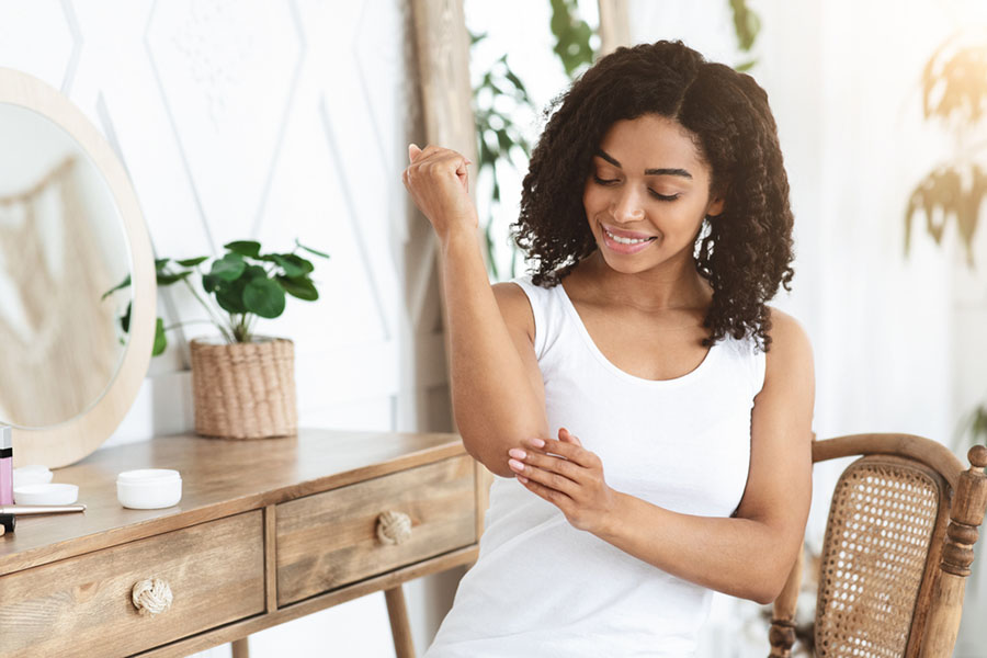 Dry Elbows Treatment. Smiling Black Woman Applying Moisturising Cream At Home, Healing Cracked Skin
