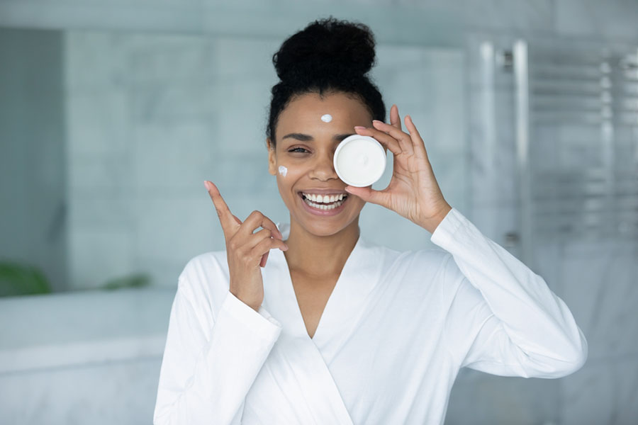 Happy beautiful young african ethnicity multiracial woman in bathrobe holding jar with cream, satisfied with professional cosmetic product results, effective daily skincare routine in bathroom.