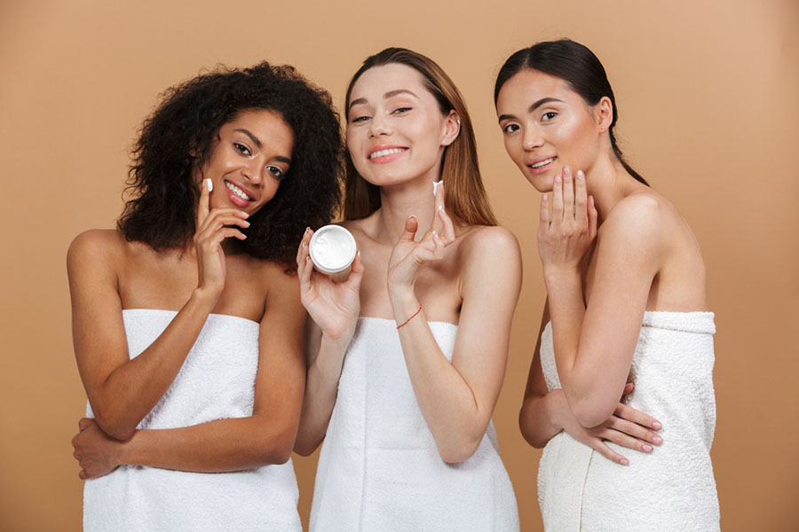 Beauty photo of three smilng multiracial women with different types of skin: caucasian, african american and asian girls, applying face cream together isolated over beige background