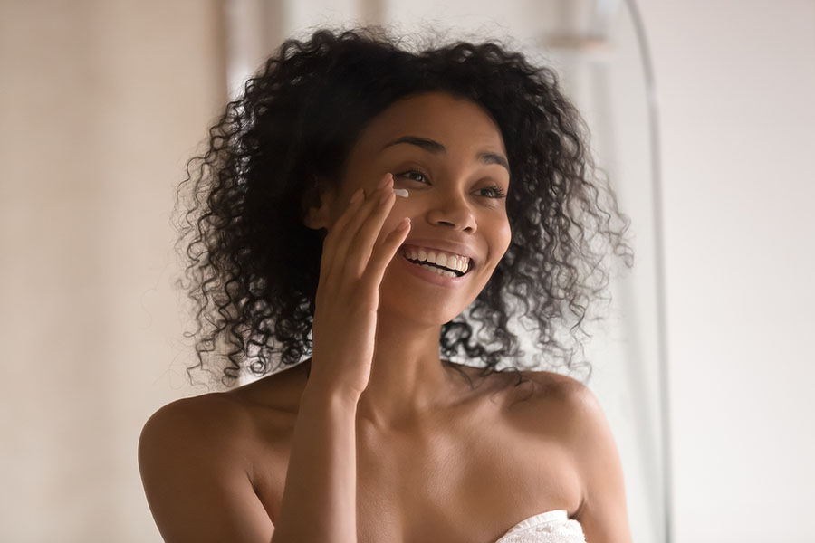 Smiling african American young woman wrapped in towel after shower look in mirror apply under eye anti-aging cream, happy biracial female perform morning skincare beauty procedures at home